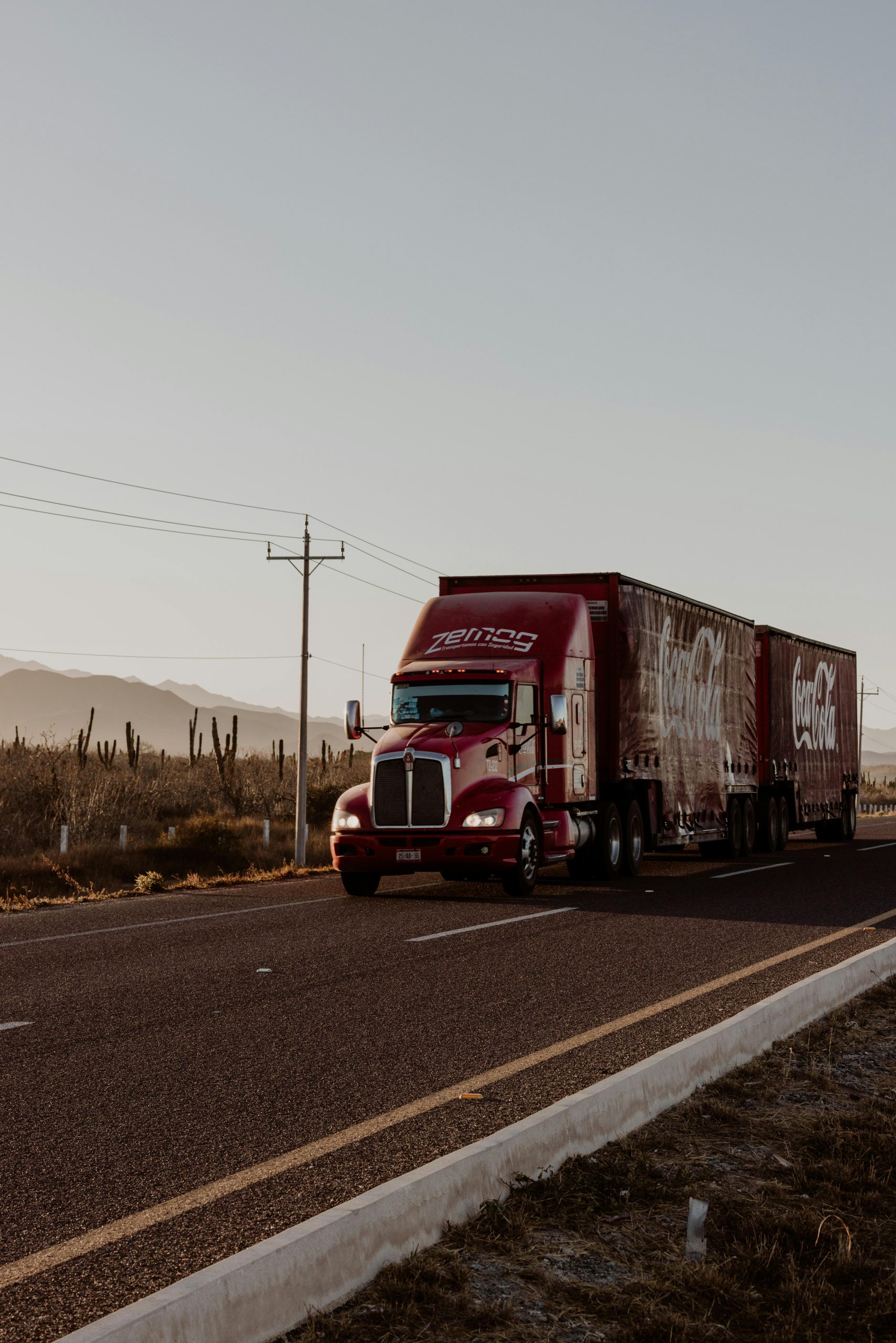 A large red truck transports goods along a desert highway under a clear sky.